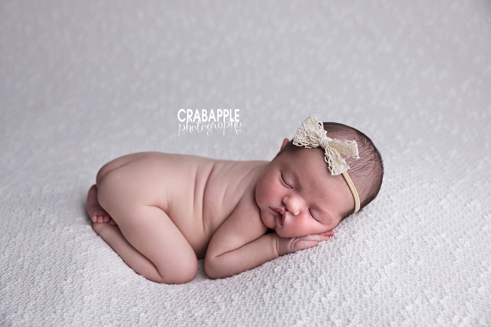 A neutral newborn baby girl photo with baby girl curled up on her tummy on a white blanket and wearing a cream colored headband