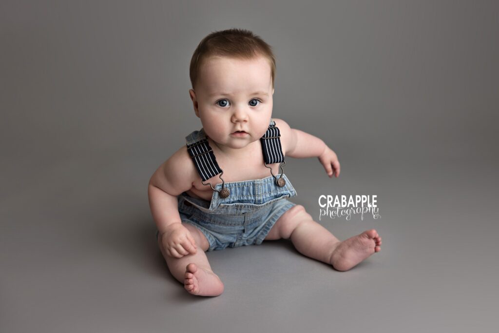 Baby boy in denim overalls sitting in front of gray backdrop sitter session baby photography 8 month photo outfit ideas