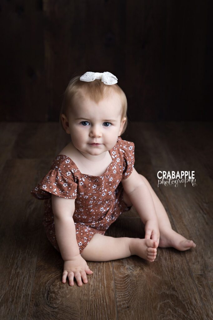 baby girl in brown floral romper sitting on dark wood floor
