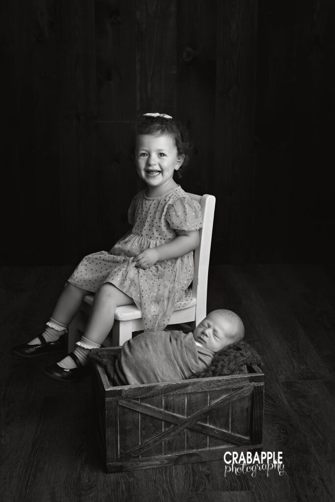 Black and white photo of toddler sister sitting on a chair next to sleeping newborn brother swaddled in a wooden crate