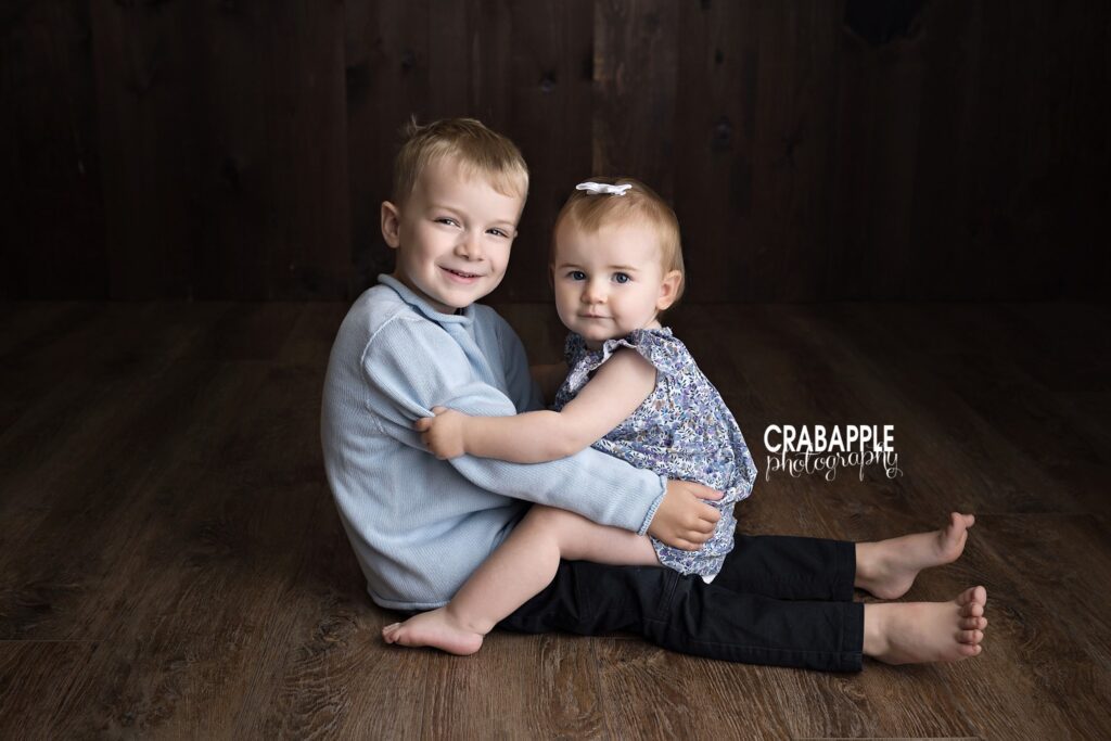 brother and baby sister sitting together smiling studio portrait

