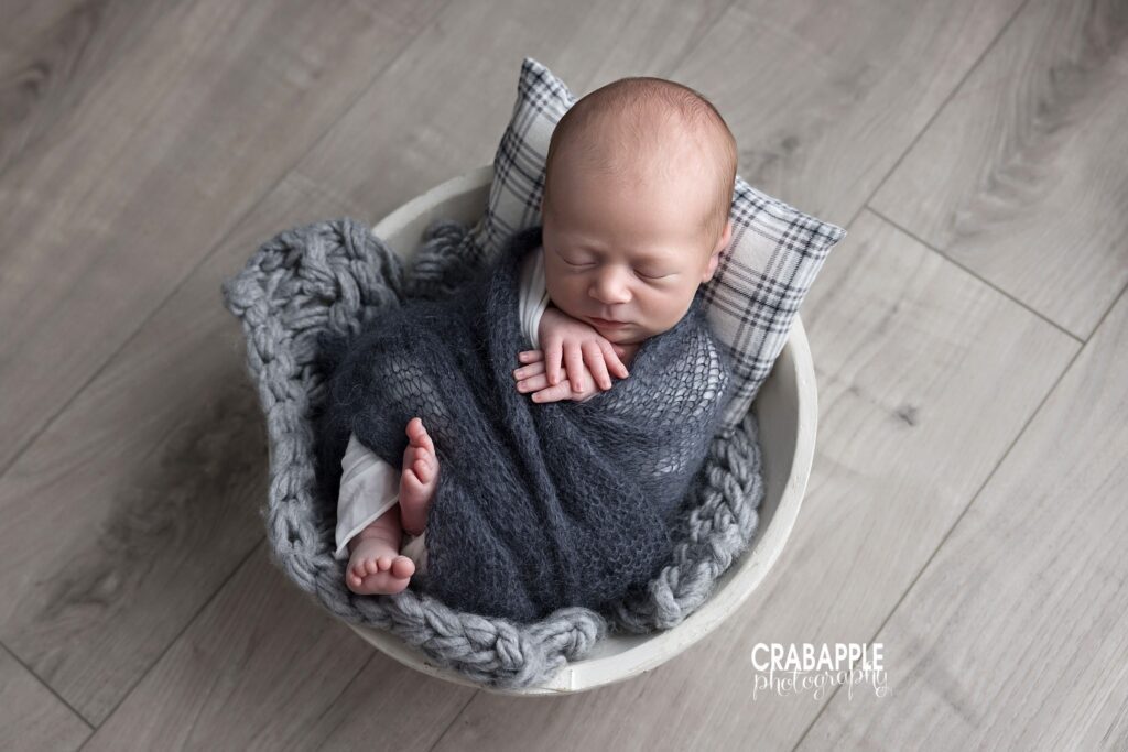 Sleeping newborn boy swaddled in gray posed in a white wooden bowl