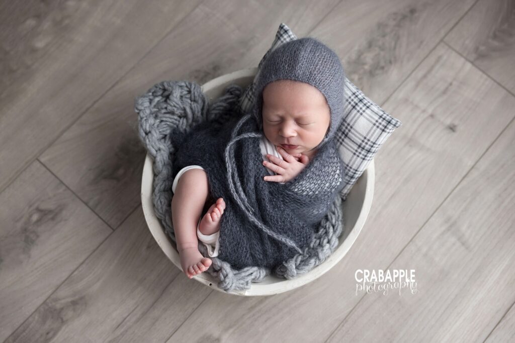 newborn baby boy posed in wooden bowl wrapped in gray
