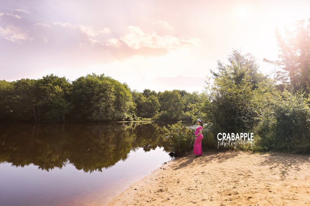 Pregnant mother in bright pink dress stands next to a pond reflecting light and trees during outdoor maternity photography session near Boston MA
