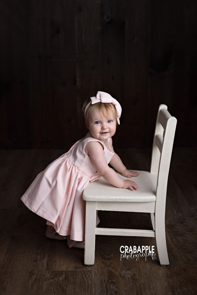 One year old girl standing beside chair in pink birthday outfit
