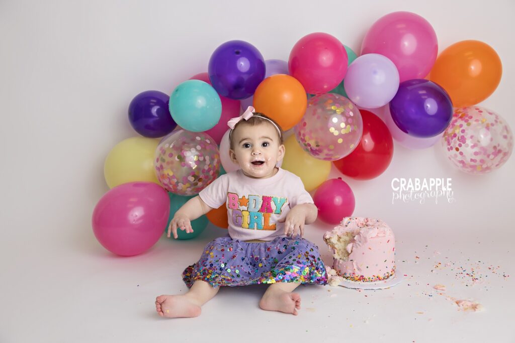 Cake Smash Setup Birthday girl sitting beside pink cake and colorful balloons