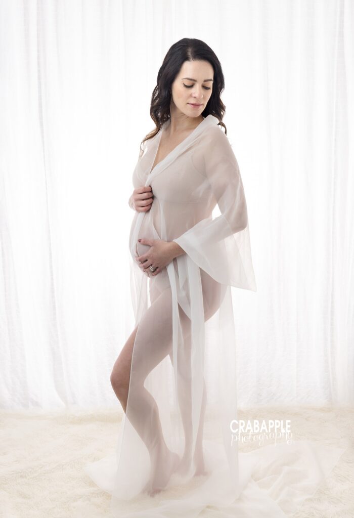 Soft studio maternity portrait Expecting mother in a sheer white robe photographed against a white curtain backdrop