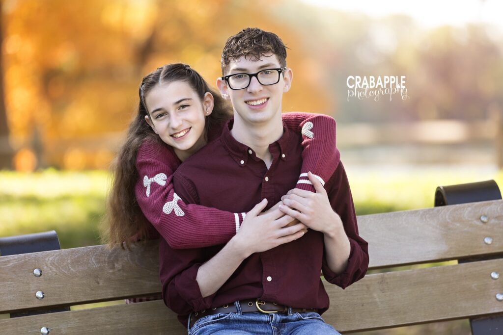 Two siblings sitting together outdoors with colorful fall foliage behind them in Medford