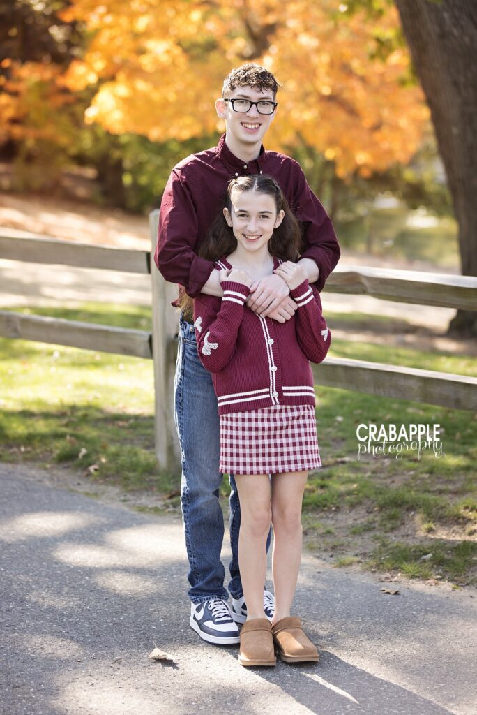 Brother and sister stand together in front of a wooden fence surrounded by fall foliage