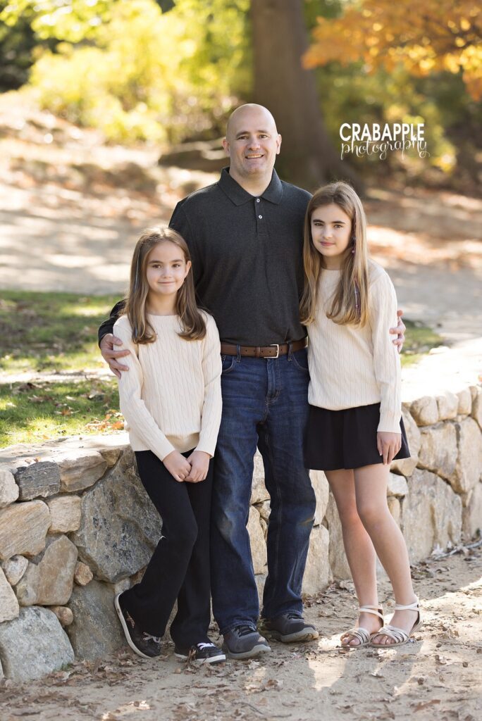 Father and daughters stand together in front of a stone wall during Medford MA family portraits in fall
