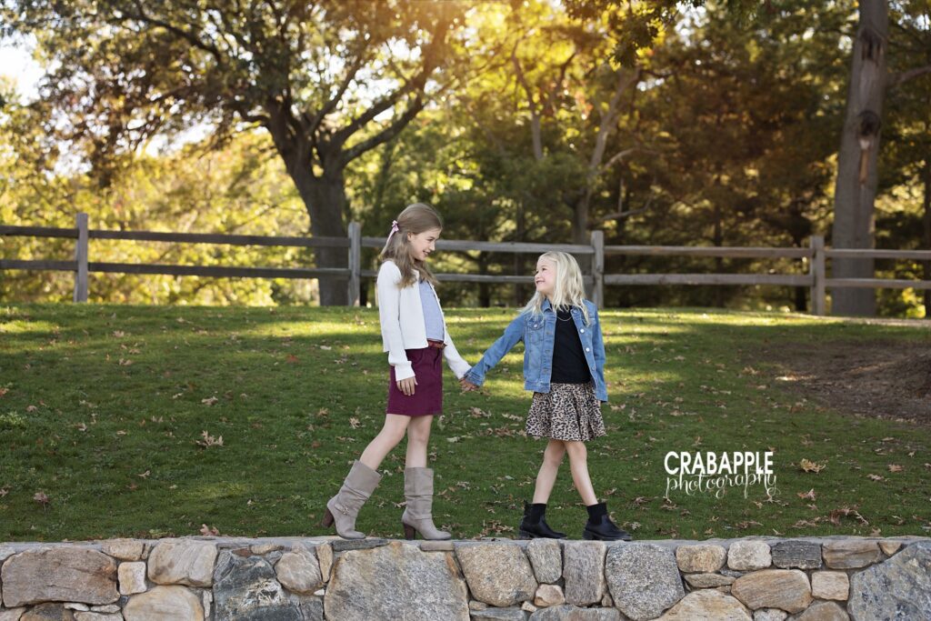 Stone wall sister photo Two sisters hold hands and walk along a stone wall during fall family photos in Medford MA