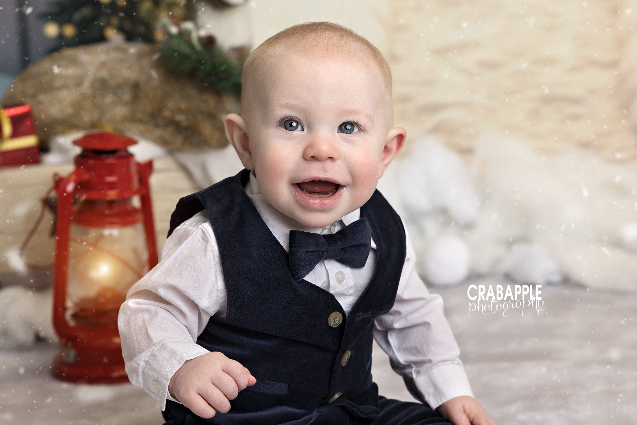 Baby boy wearing a dark bow tie and suspenders posing on a light blanket beside Christmas decorations	