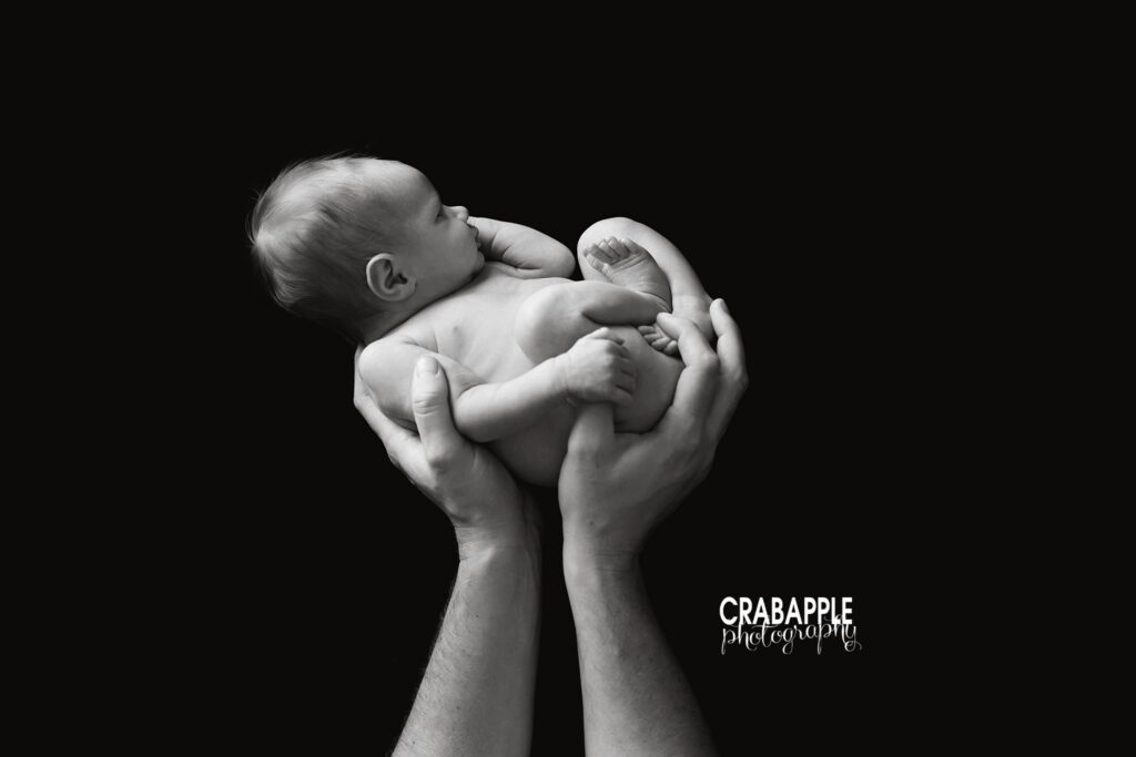 Black-and-white portrait of a newborn baby curled in her father's hands against a black background
