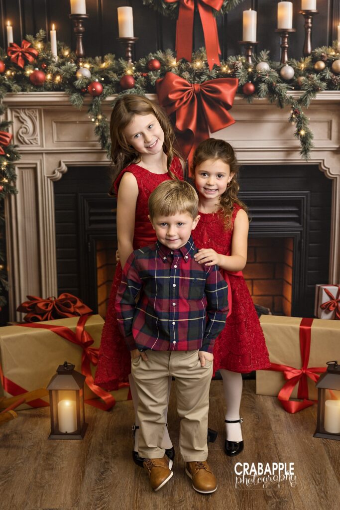 Three siblings posing together in front of a roaring fireplace set design for Christmas portraits
