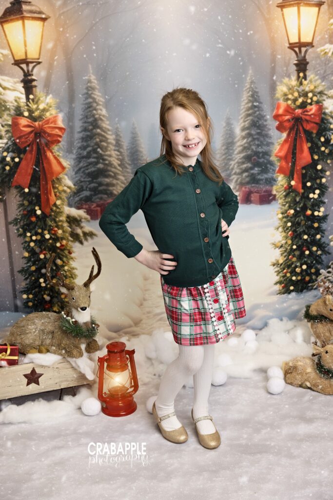 Young girl in a plaid holiday skirt and sweater posing in front of a decorated Christmas scene