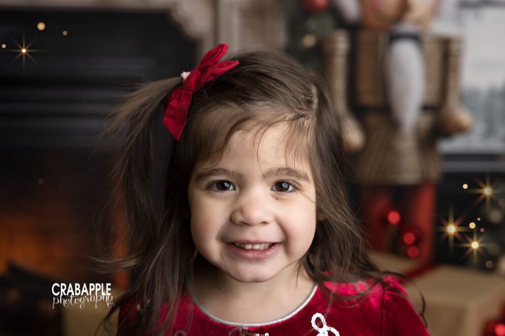 Smiling young girl in a red holiday dress posing in front of a decorated Christmas set