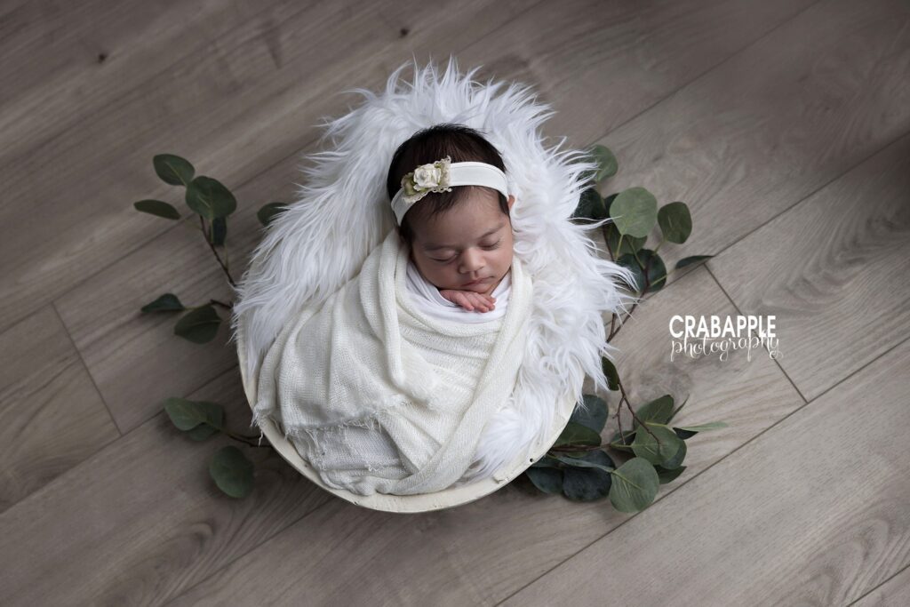 White-swaddled newborn girl in a fur-lined white wooden bowl surrounded by faux greenery	