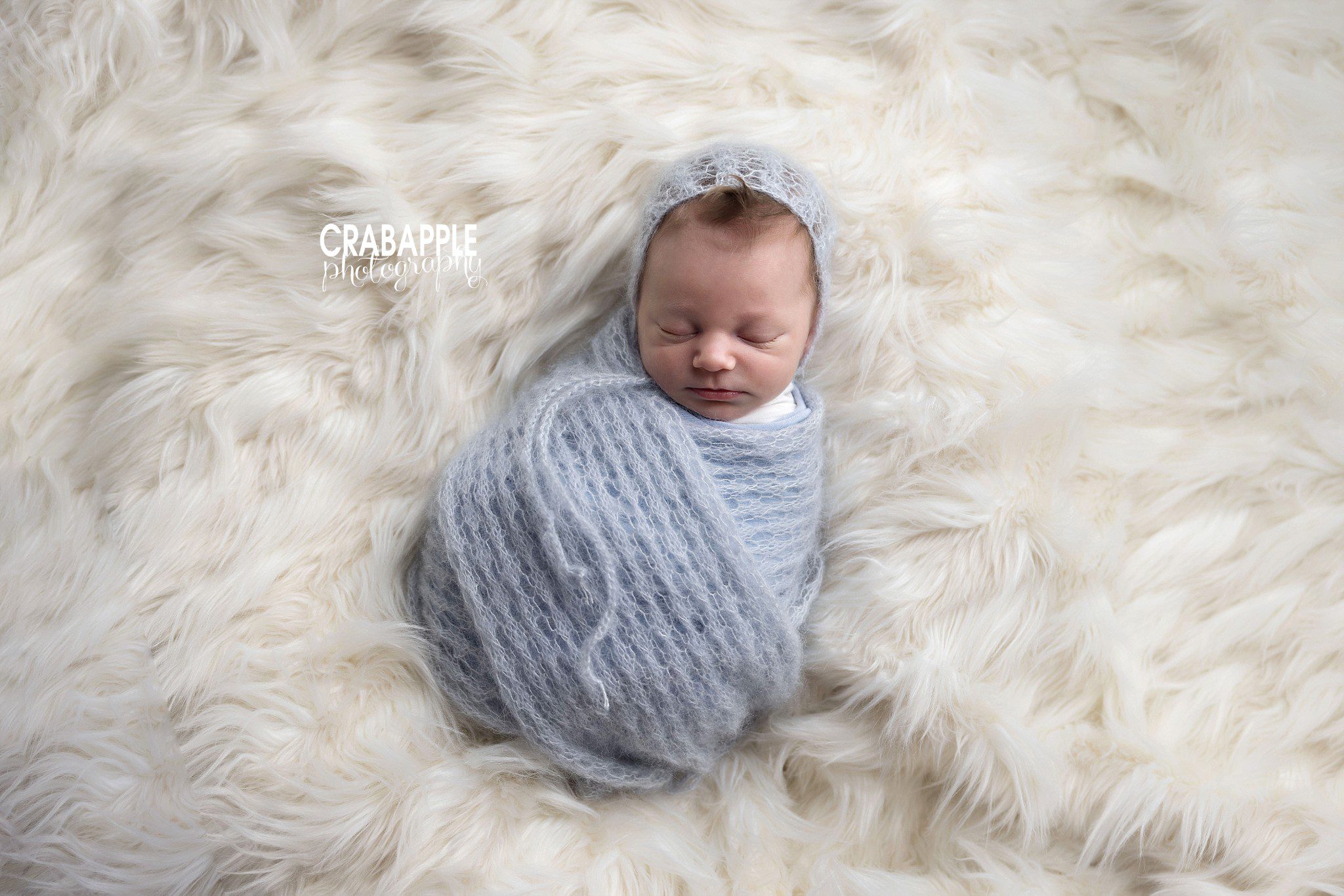 Newborn baby boy peacefully swaddled in blue on soft white fur

