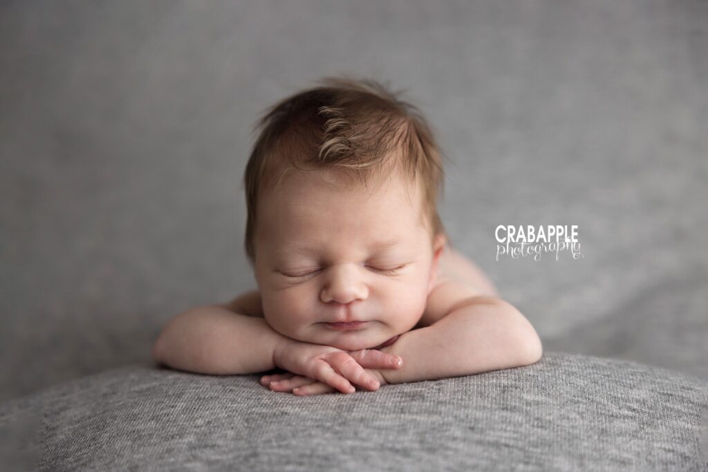 Newborn baby boy resting his chin on his hands on a gray blanket
