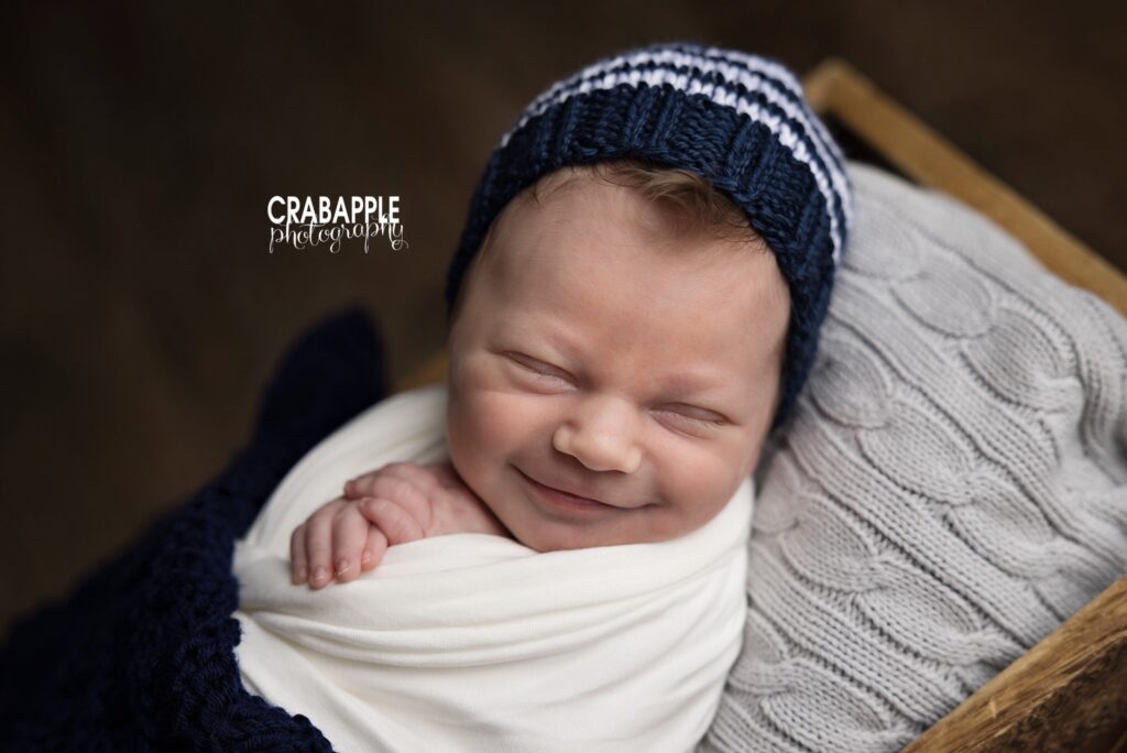 Smiling newborn baby boy in a white swaddle resting in a wooden crate
