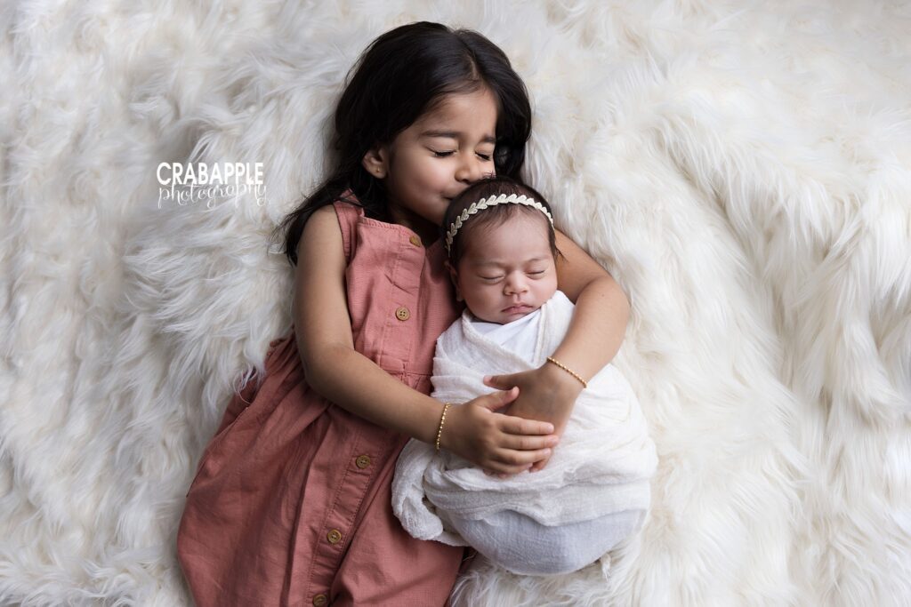 Big sister gently kissing her newborn baby sister, who is swaddled in white	