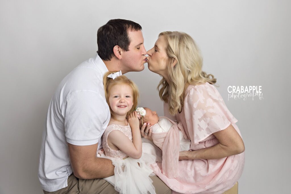 Mom and dad kissing while their toddler daughter smiles at the camera and their newborn is held close
