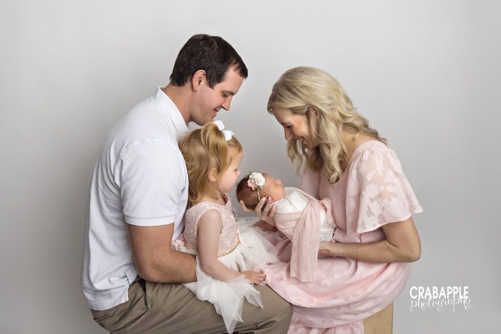 Dad holding his toddler daughter while mom cradles their newborn and the parents look toward each other
