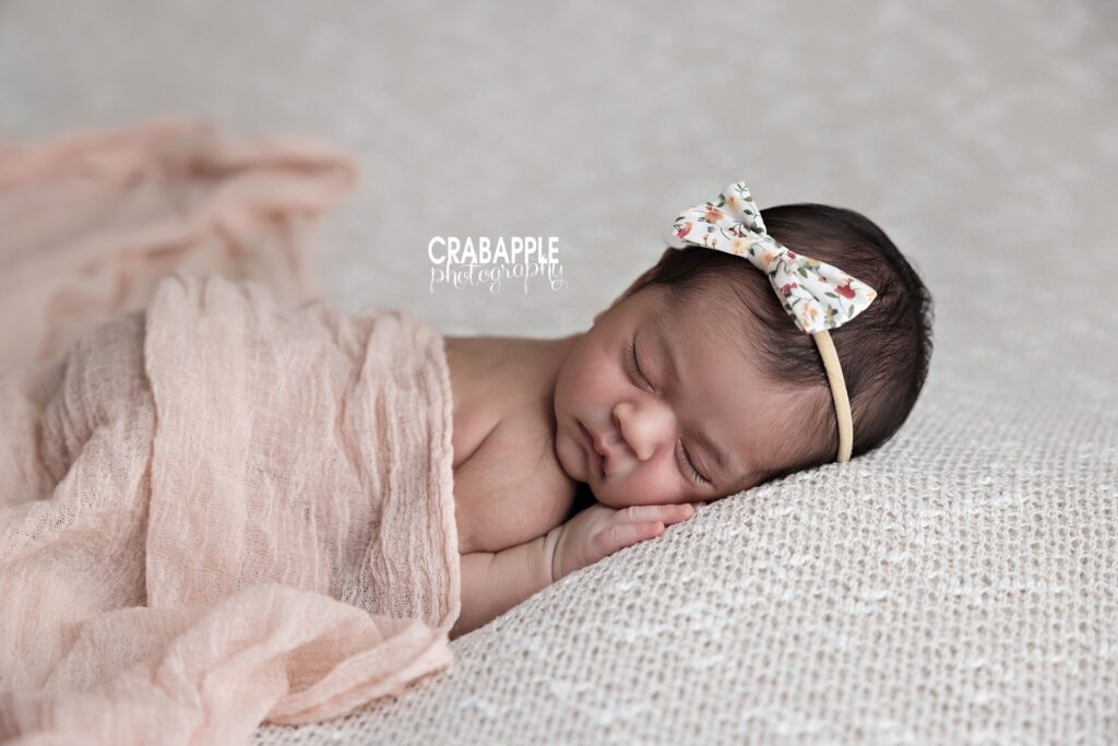 Newborn girl lying on her stomach with her head turned to the side, wearing a delicate floral headband and blush swaddle	