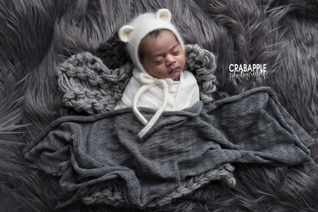 Newborn girl wearing a white bear hat and white swaddle against gray fur and chunky knit textures	