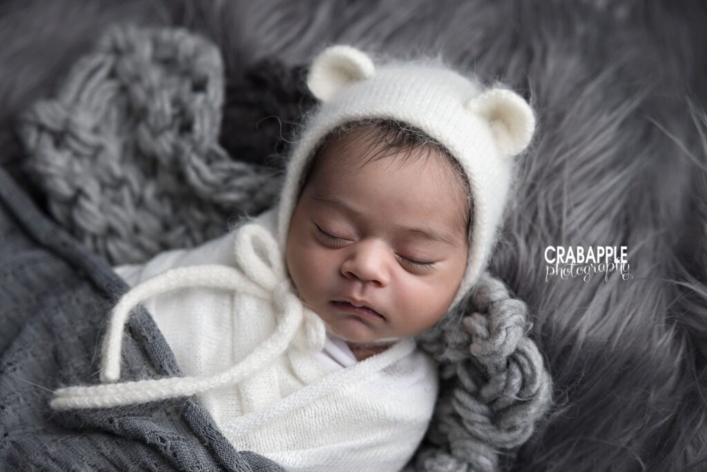 Close-up of newborn girl wearing a white bear hat and white swaddle on gray chunky knit and soft fur	