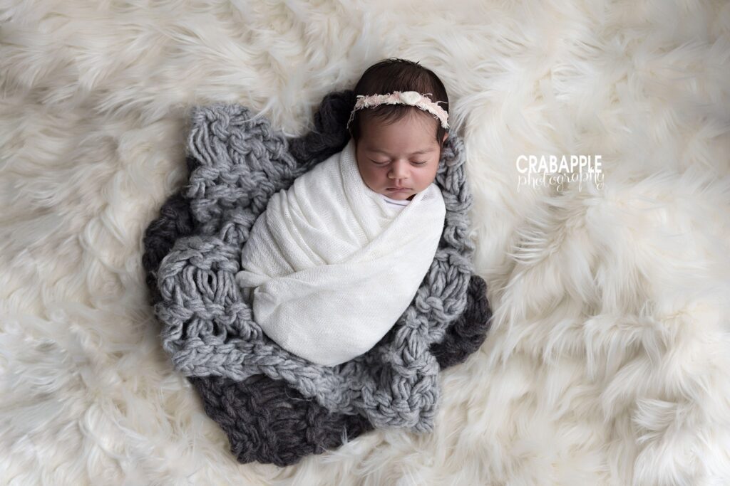 Newborn girl in a white swaddle and delicate headband resting on gray knit fabric and white fur	