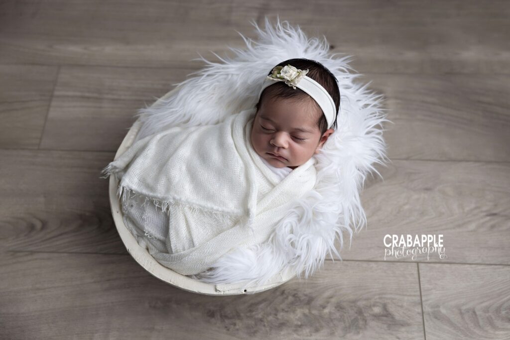 Newborn girl swaddled in white and resting in a white wooden bowl lined with soft white fur	