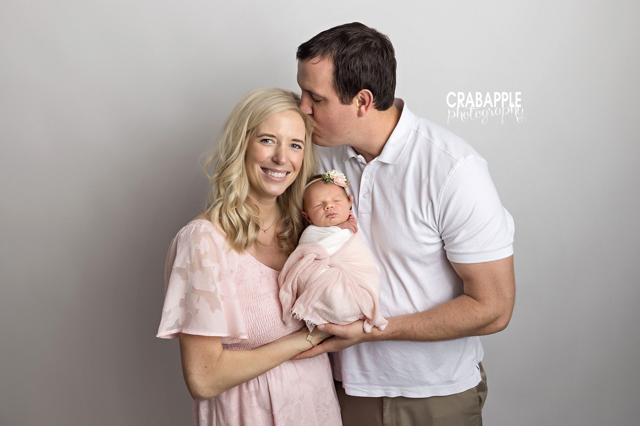 Mom and dad holding their newborn baby together as dad kisses the top of mom's head
