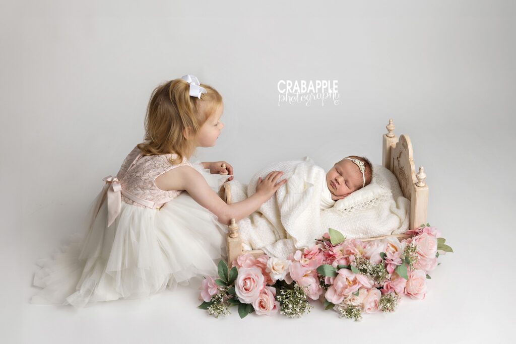 Big sister looking lovingly at her sleeping baby sister in a small bed decorated with flowers
