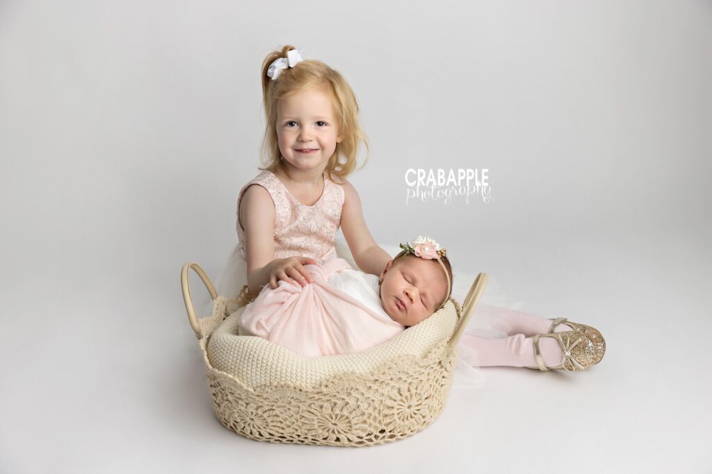 Sleeping newborn baby girl wrapped in pink in a crochet-style basket with her big sister sitting behind her

