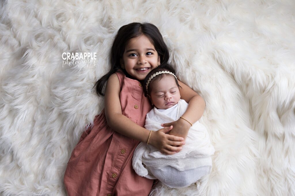 Three-year-old big sister in pink smiling beside her newborn sister wrapped in white	