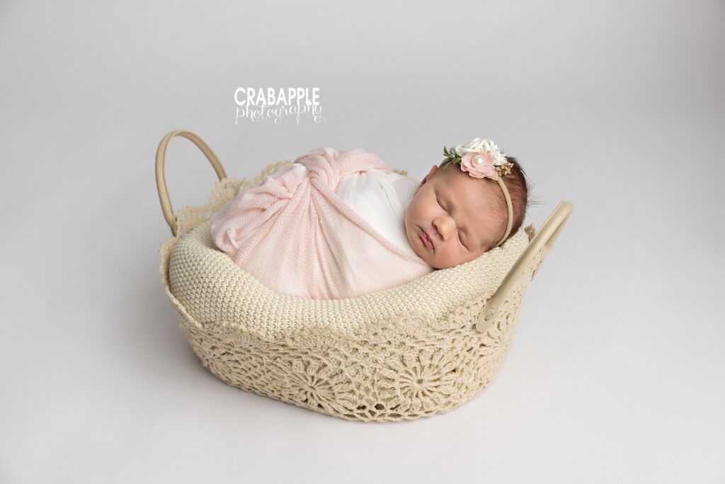 Sleeping newborn baby girl wrapped in white in a crochet-style basket with a delicate floral headband