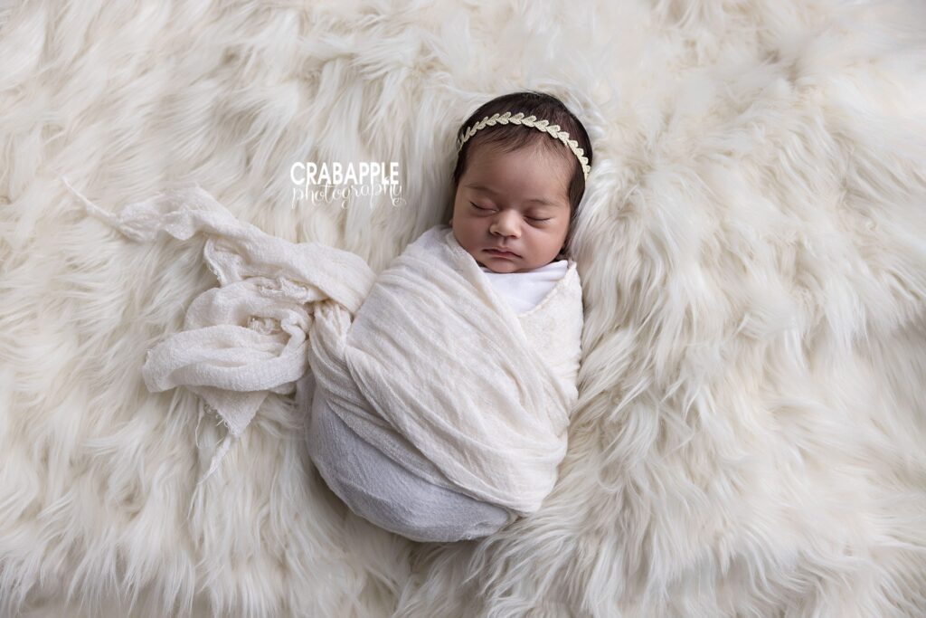 Newborn girl wrapped in a white swaddle and photographed on soft white fur	